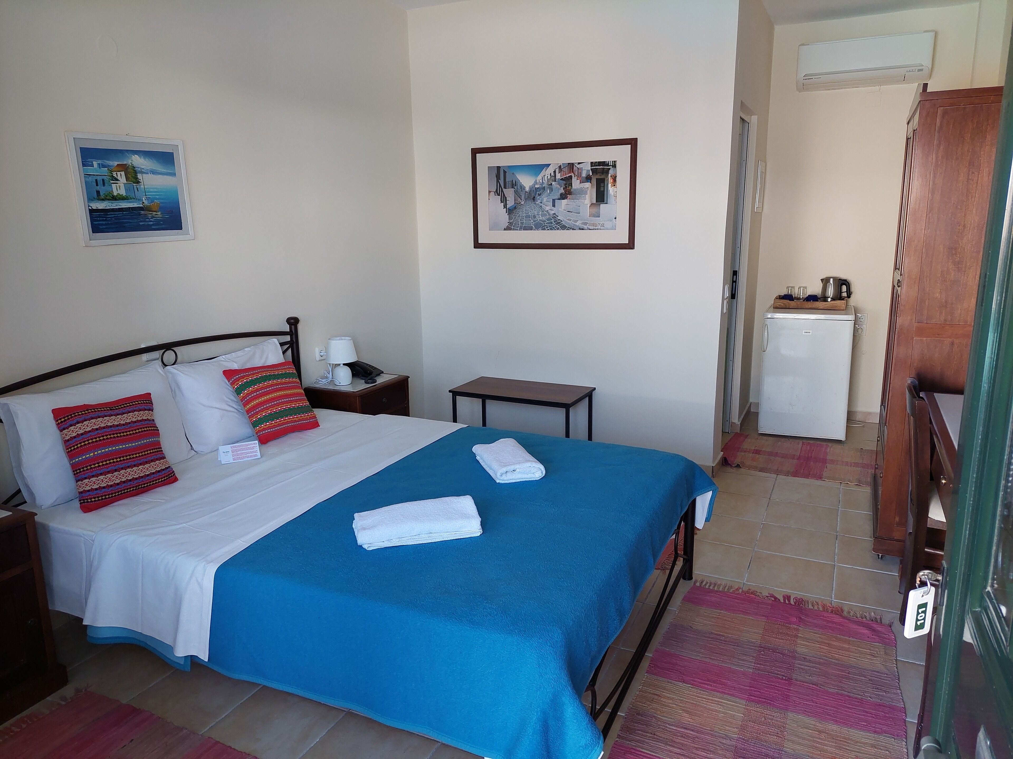 Una cama bien hecha con un edredón azul y dos almohadas, una mesita de noche con una lámpara, aire acondicionado en la pared y un refrigerador en la esquina.