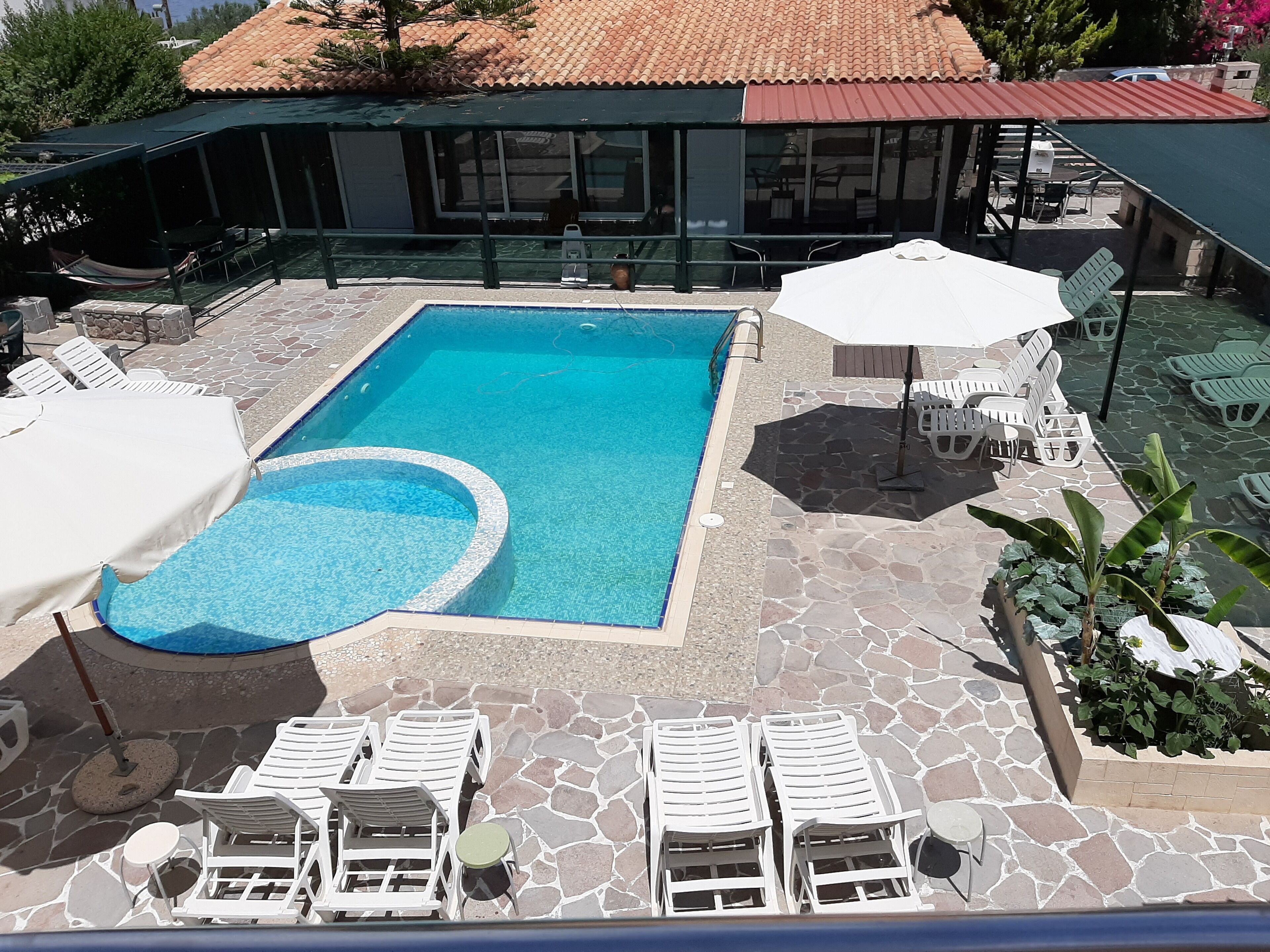 A swimming pool with lounge chairs and umbrellas, surrounded by stone tiles and a covered area with chairs and tables.