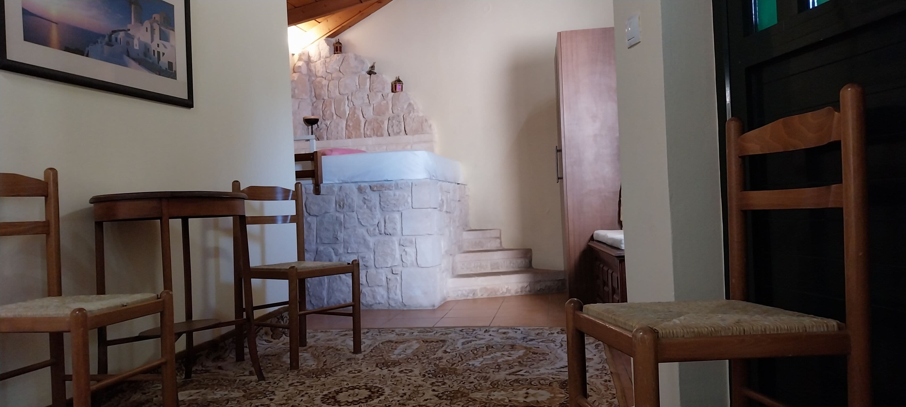 Una habitación con pared de piedra, una cama, una silla de madera, una mesita y una puerta.
