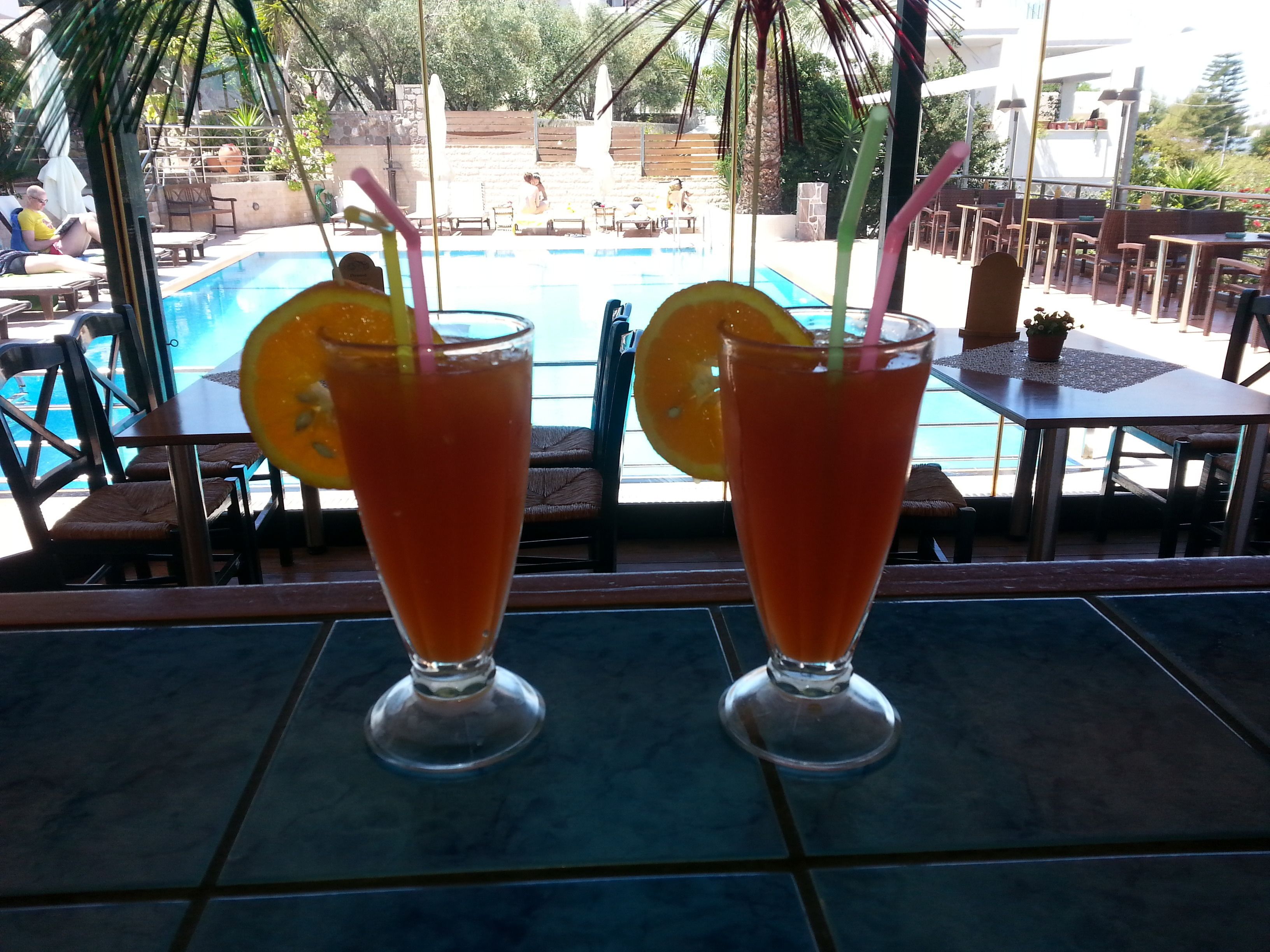 Two tall glasses of orange juice with straws and orange slices on a tiled table near a poolside area with lounge chairs and umbrellas.