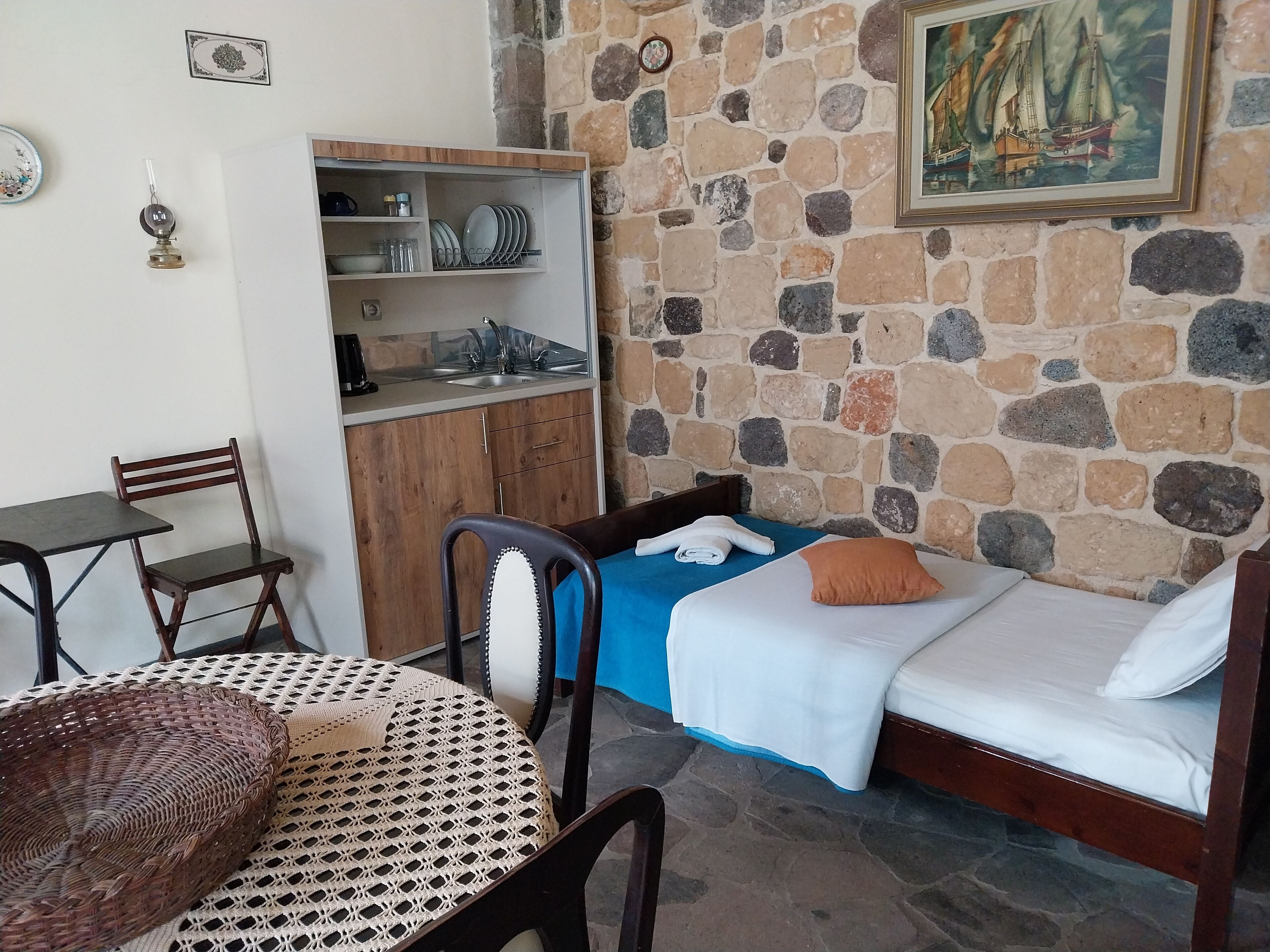 Une pièce avec un mur en pierre, un lit recouvert d’une couverture bleue et orné d’un coussin orange, une armoire en bois, une table à manger avec un panier tressé, et une chaise.