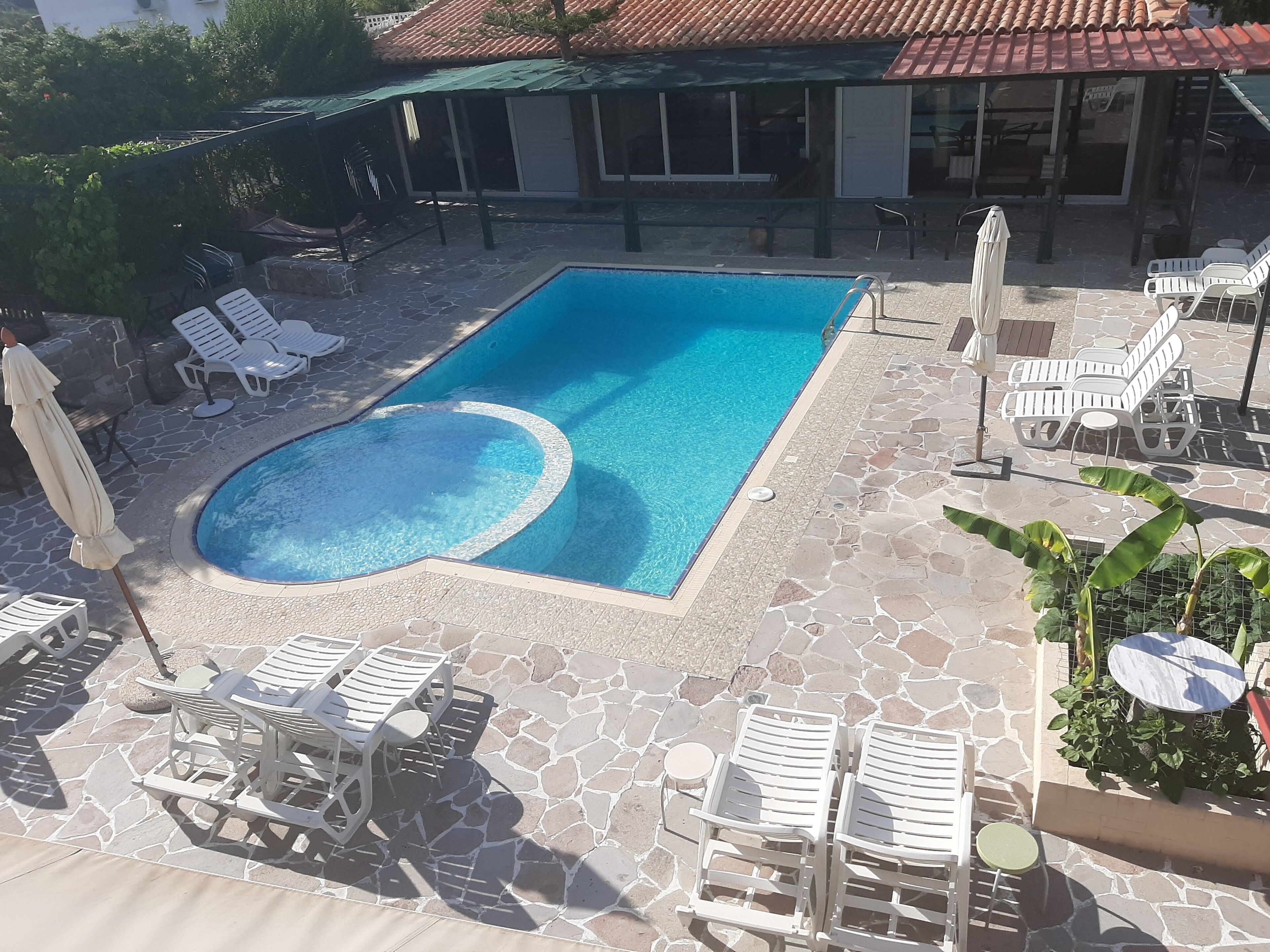 A swimming pool with lounge chairs and umbrellas, surrounded by stone tiles and a covered area with a tiled roof.