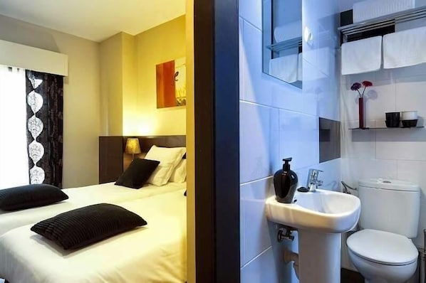 A hotel room with a bed, a bedside table, a lamp, a window with curtains, and a bathroom with a sink and toilet. - THC Tirso Molina Hostel (Madrid)