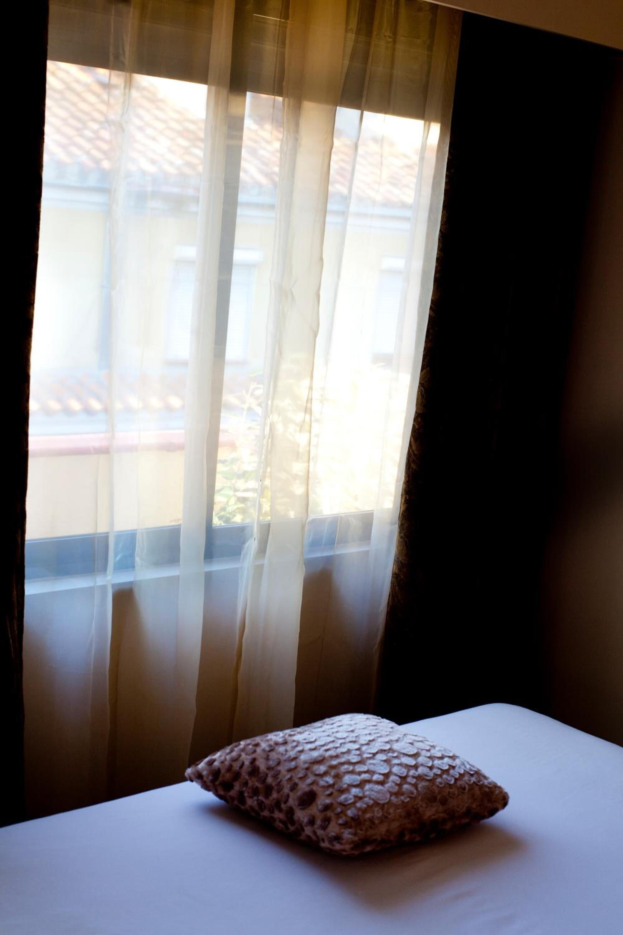 Cama com travesseiro estampado e cortinas leves entreabertas, revelando a vista de edifícios lá fora.