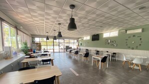 Un vaste espace dédié à la salle à manger, avec de nombreuses tables et chaises, de grandes fenêtres laissant entrer la lumière naturelle et un mur végétal agrémenté d’une horloge.