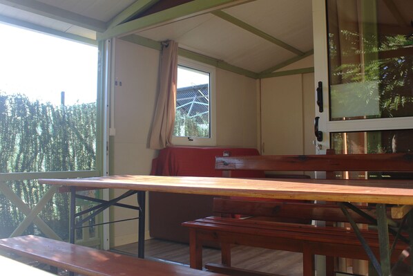 Une pièce avec une table et des bancs en bois, une fenêtre avec un rideau et une vue sur les arbres à l’extérieur.