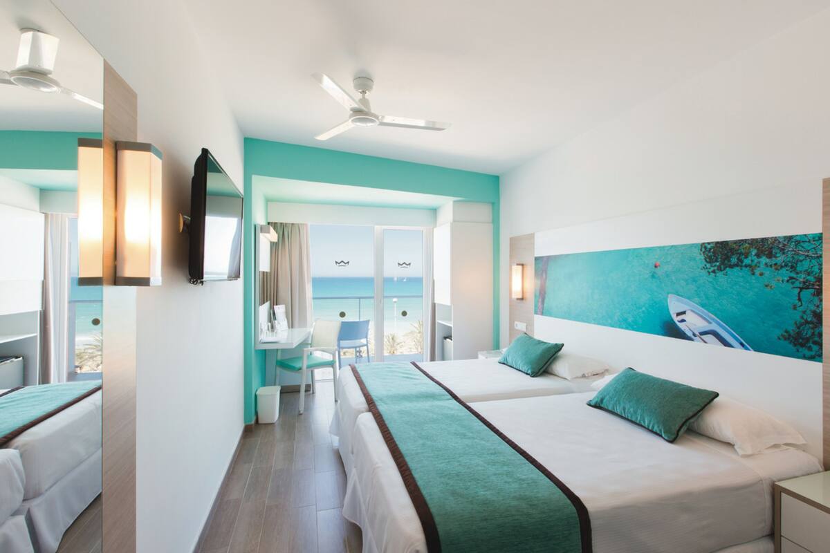 A hotel room with two beds, a desk, a chair, and a large window with a view of the sea.
