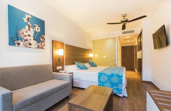 A hotel room with a sofa, a bed with a blue floral bedspread, a wooden headboard, a ceiling fan, a flat-screen TV, and a painting of flowers on the wall.