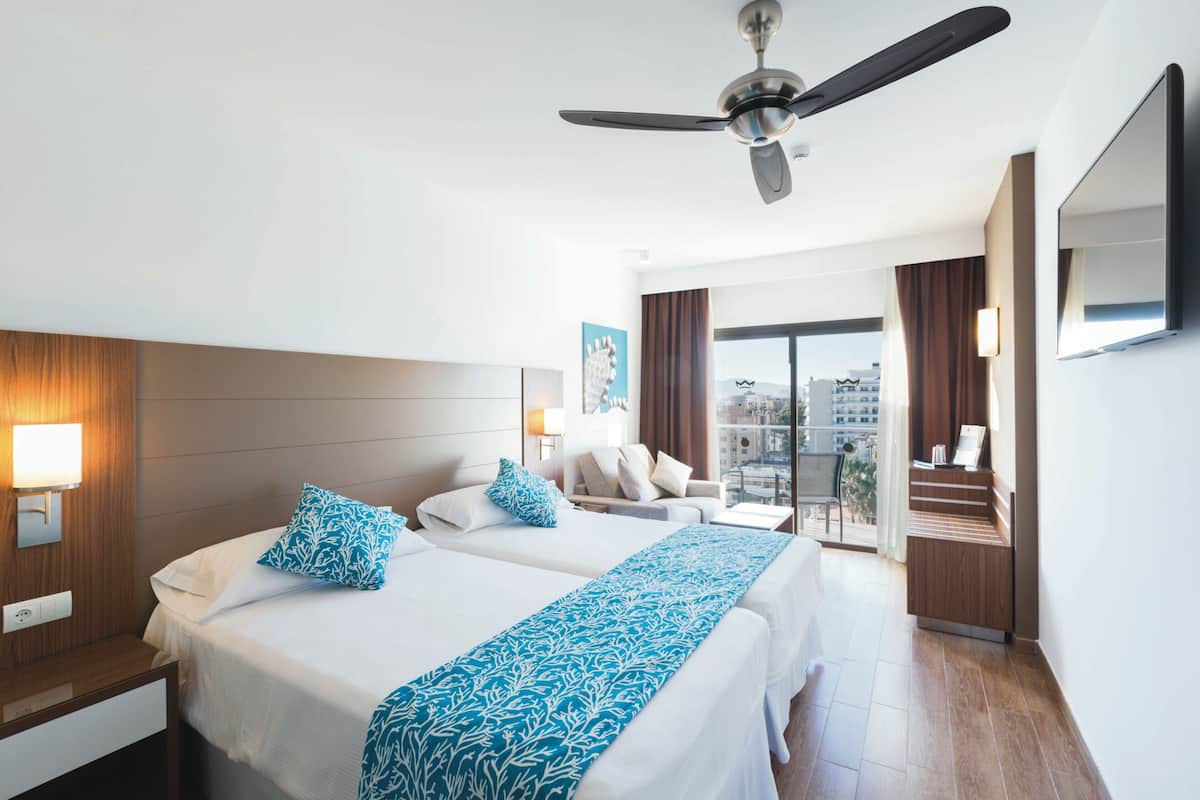 A hotel room with two beds, a ceiling fan, a balcony with a view of buildings, and a TV mounted on the wall.