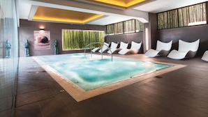 Un espace piscine intérieur moderne, doté d’une grande piscine, d’un coin salon avec des chaises blanches et d’un mur orné de lattes de bois verticales.