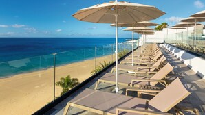 A beachside balcony with lounge chairs and umbrellas, overlooking a sandy beach and the ocean.