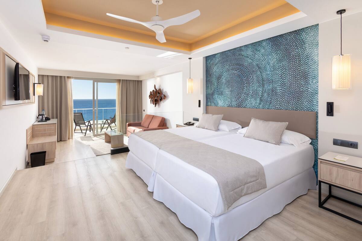 Una habitación de hotel moderna con una cama grande, un ventilador de techo, vista al océano y un espacio de descanso con una silla y una mesa.