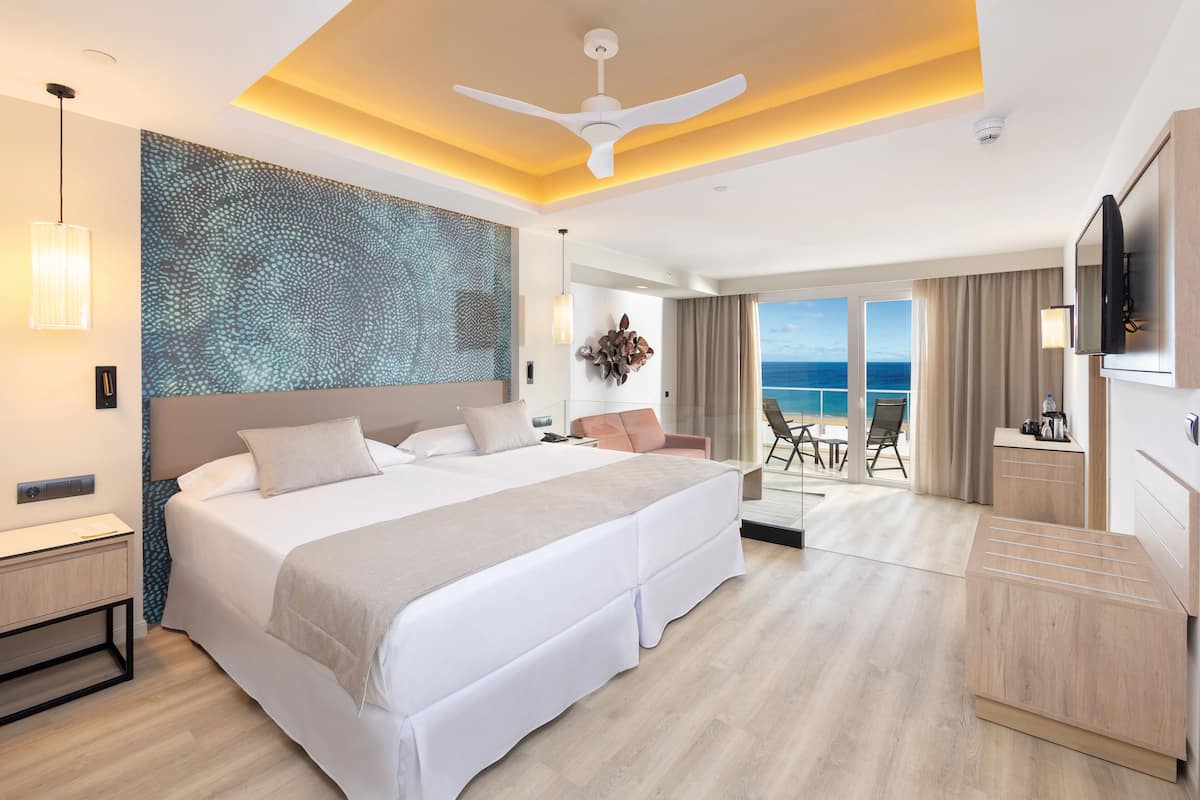 Una habitación de hotel moderna con una cama grande, un ventilador de techo, vista al océano y un balcón con mesa y sillas.