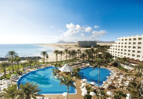 A resort with two large swimming pools, a beach, and a hotel building.