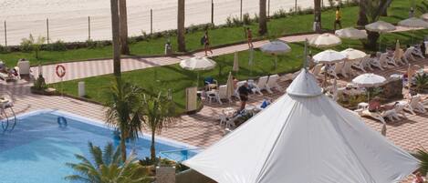 A resort pool area with lounge chairs and umbrellas, surrounded by palm trees and a beach in the background.