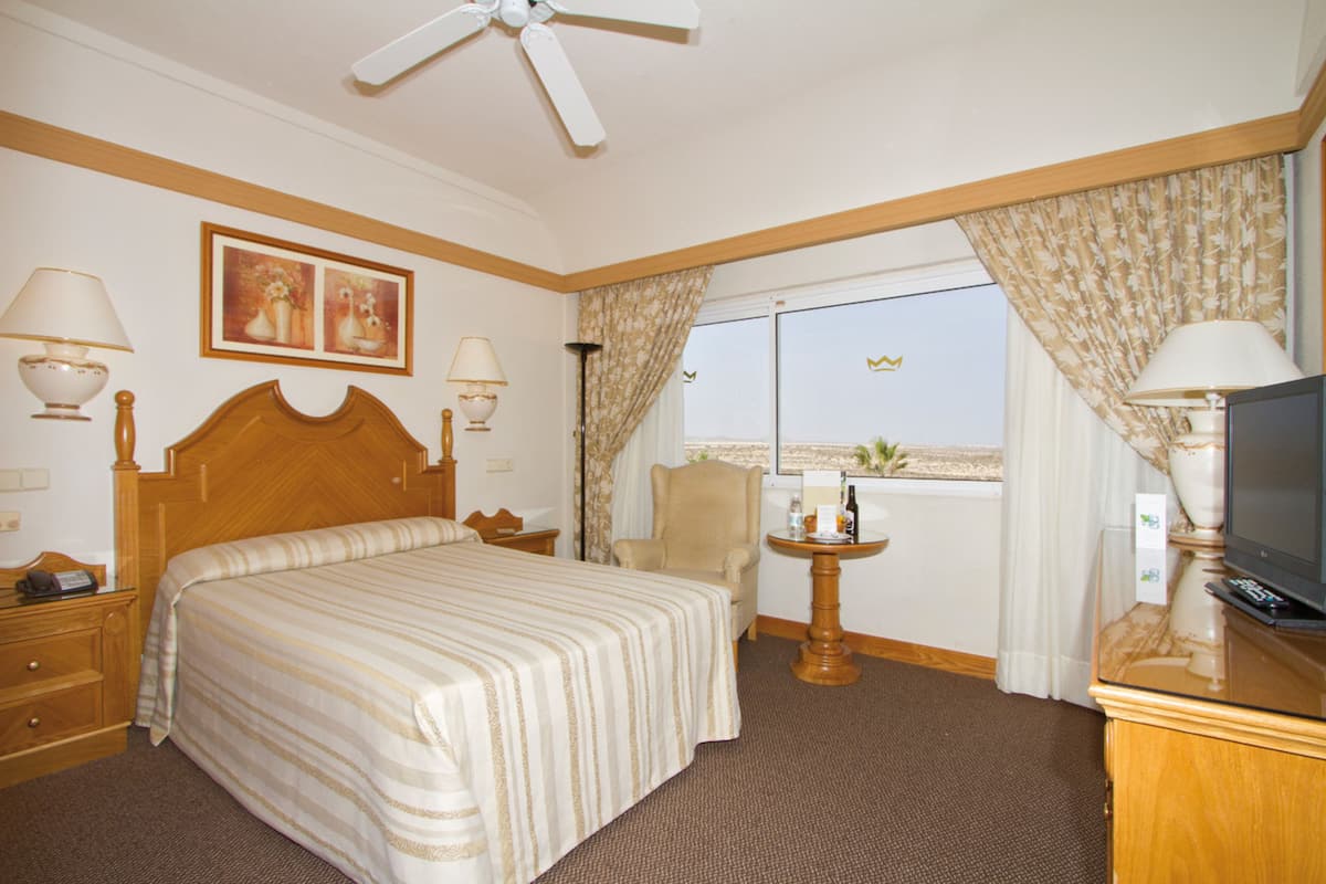 A hotel room with a large bed, a television, a desk with a lamp, and a view of the beach.