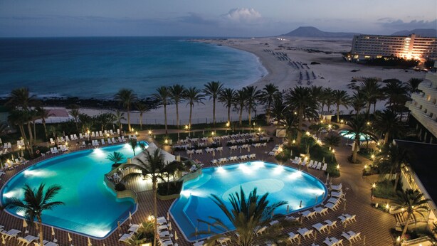 A resort with two large swimming pools surrounded by lounge chairs and palm trees, overlooking a beach and ocean. - Hotel Riu Palace Tres Islas (La Oliva)