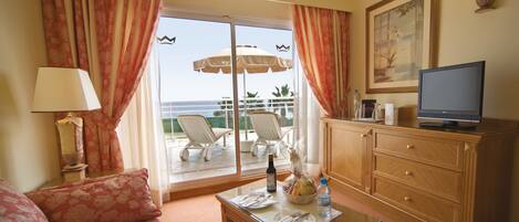 A hotel room with a sofa, a television on a wooden cabinet, a dining table with a bottle and glasses, and a balcony with chairs and an umbrella.