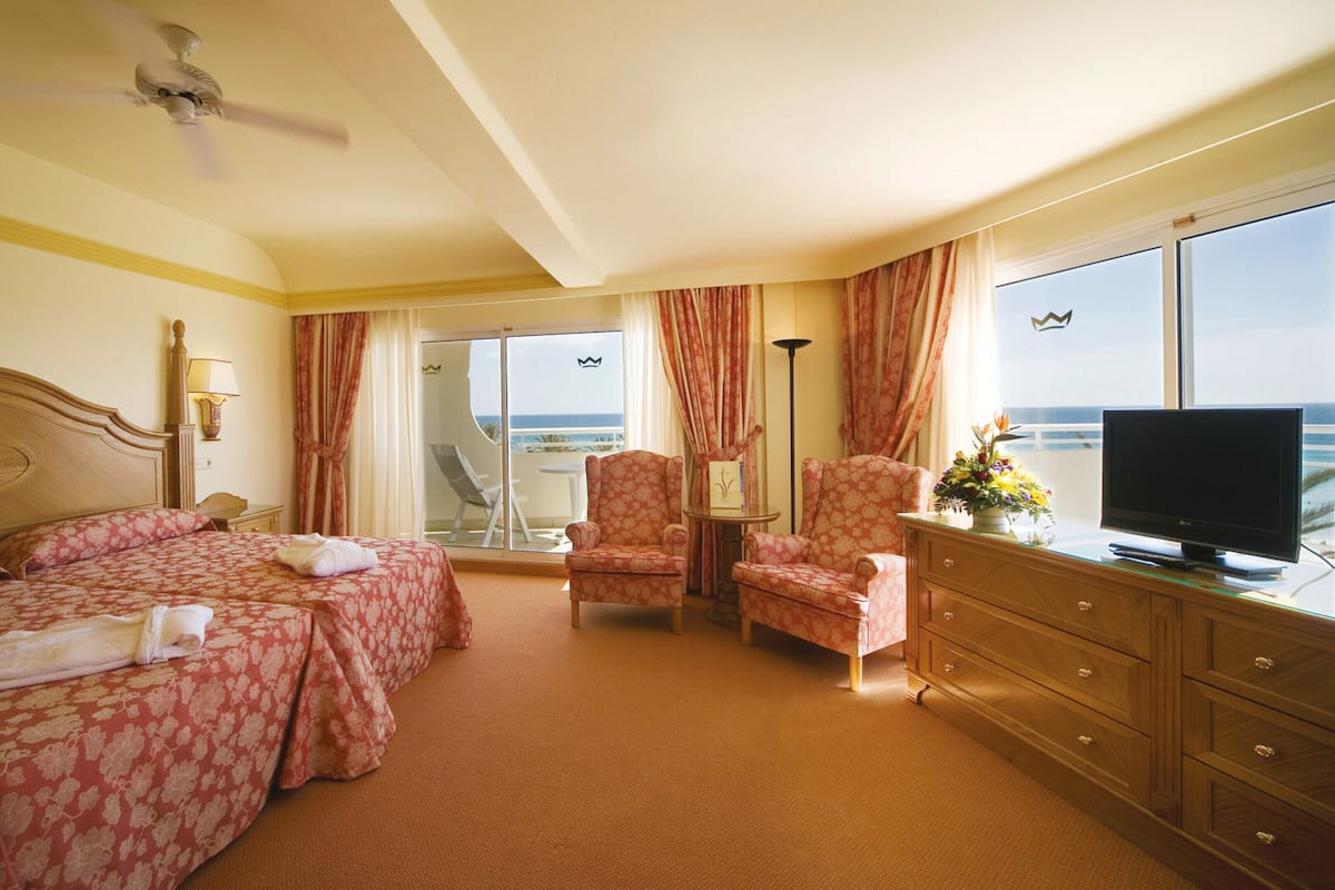 A hotel room with a bed, two chairs, a TV, a dresser, and a view of the ocean.