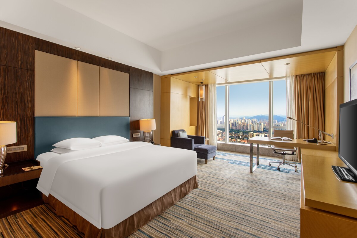 A hotel room with a large bed, a desk, a chair, and a view of a cityscape.