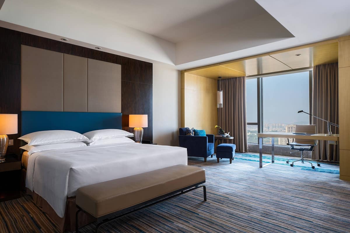 A hotel room with a large bed, a desk, a chair, and a view of the city through large windows.