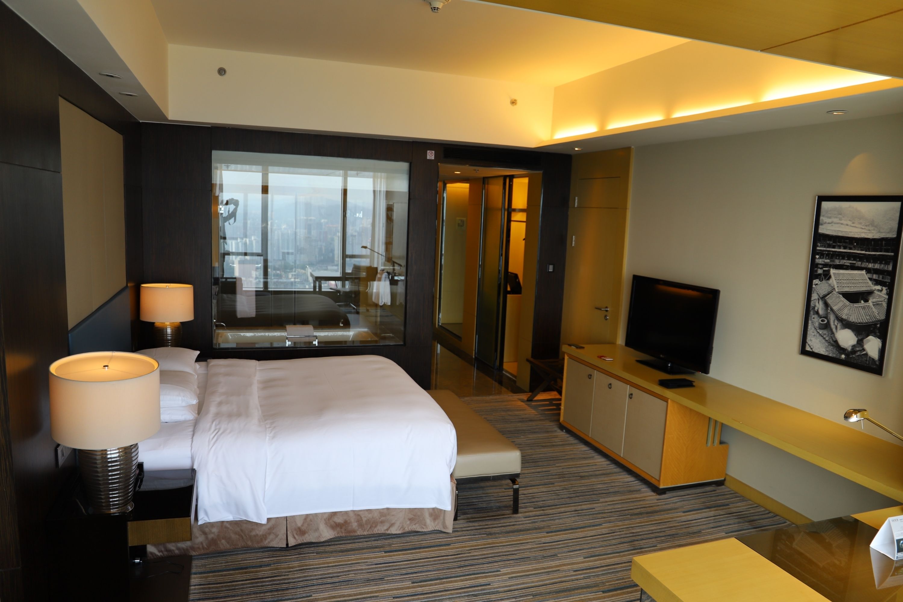 A modern hotel room with a large bed, a TV, a desk, and a view of the city.