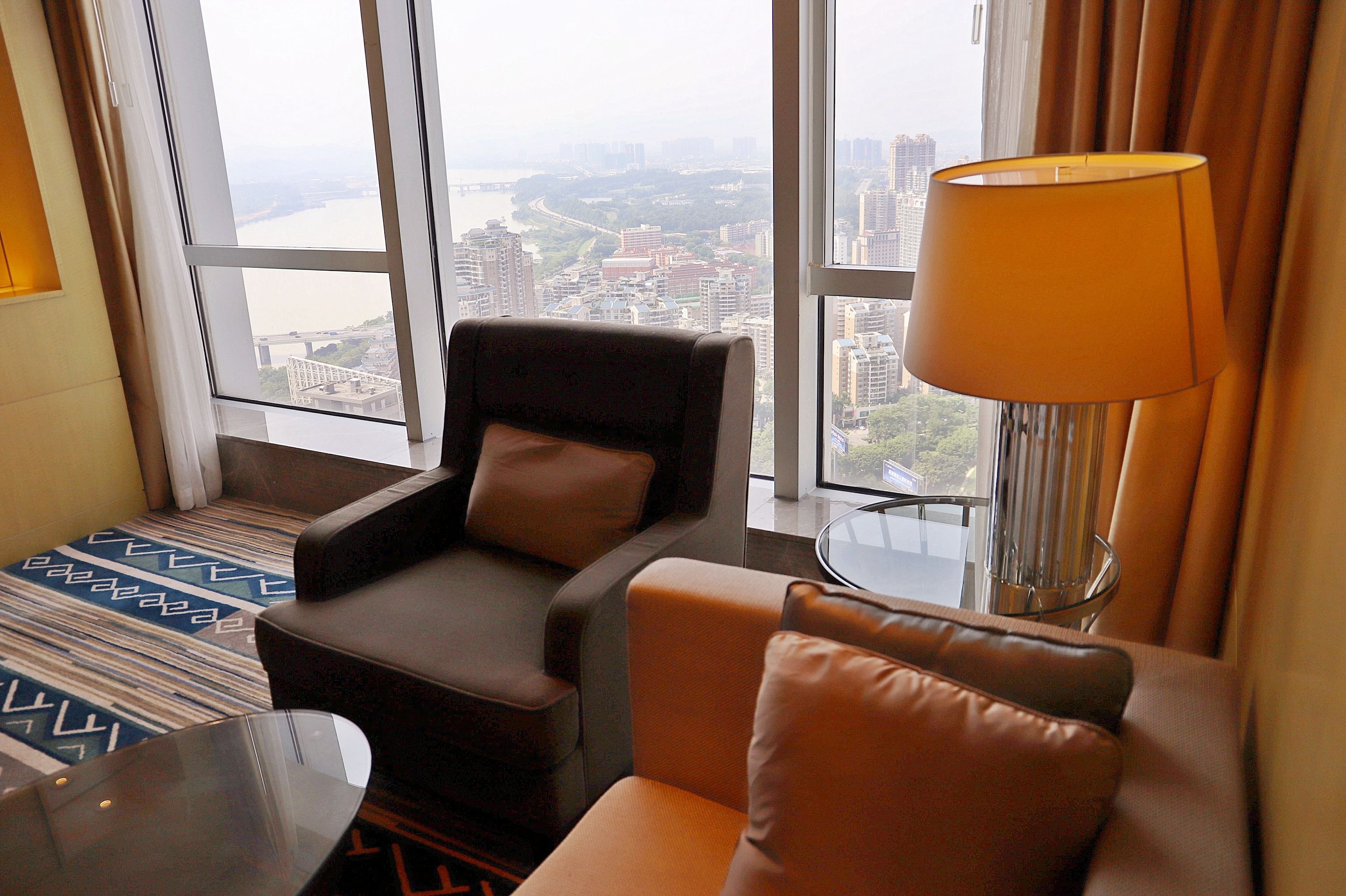 A hotel room with a large window offering a city view, a sofa, a chair, and a lamp.