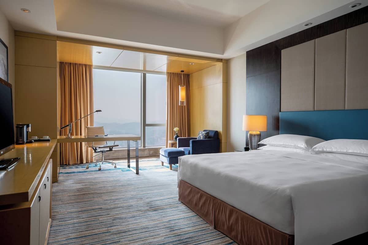 A hotel room with a large bed, a desk with a computer, a chair, and a view of the city through the window.