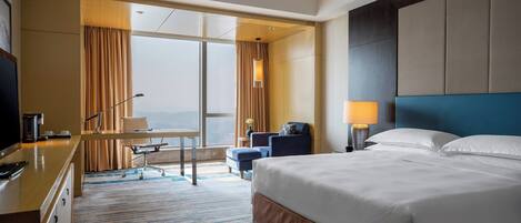 A hotel room with a large bed, a desk with a computer, a chair, and a view of the city through the window.