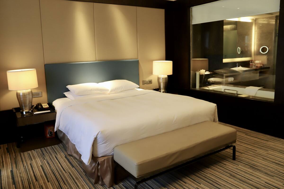 A hotel room with a large bed, two bedside lamps, a nightstand, a bench, and a large mirror.