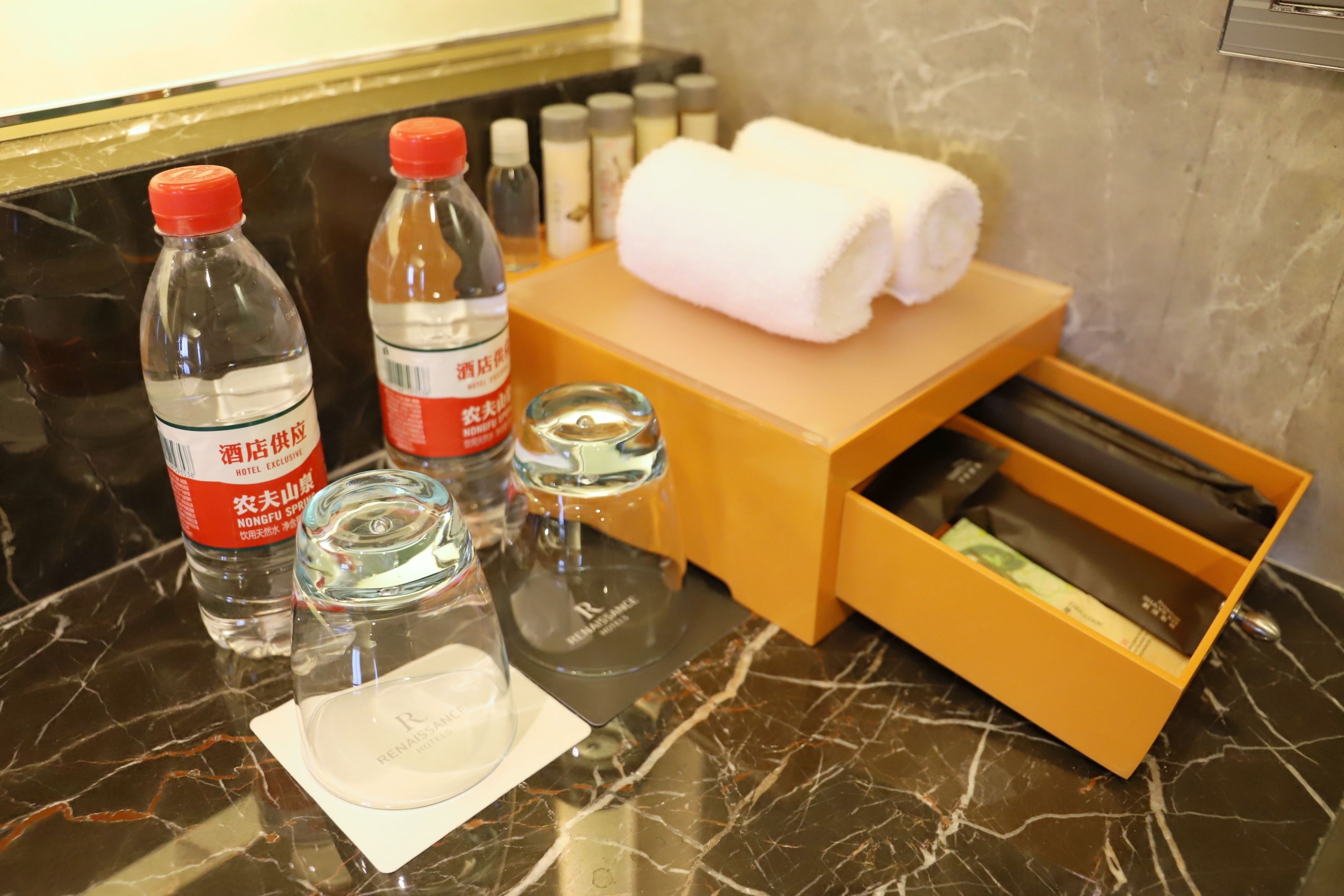 A hotel minibar with bottled water, a glass, a towel, and a box of toiletries.