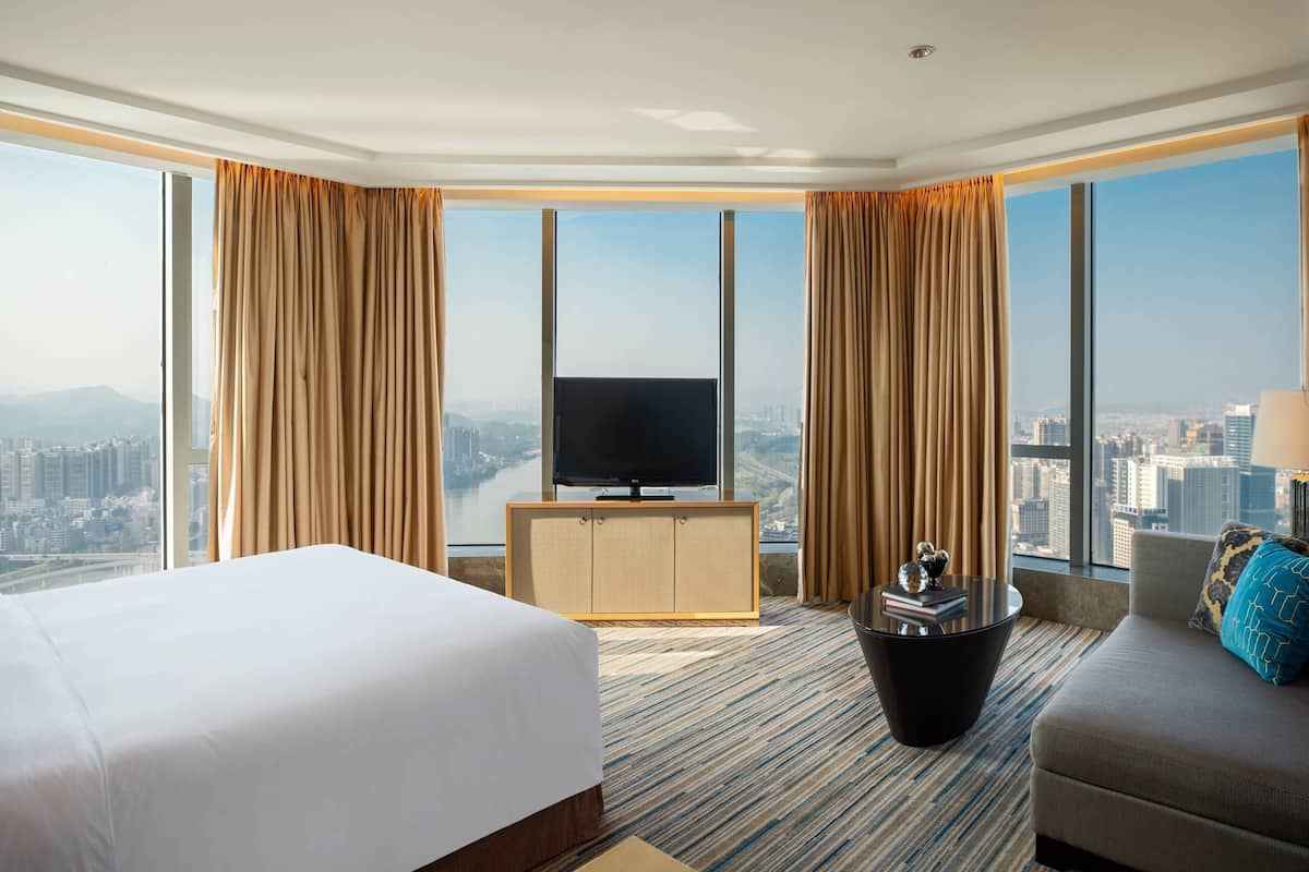 A hotel room with a bed, a sofa, a TV on a wooden cabinet, and a view of the city.