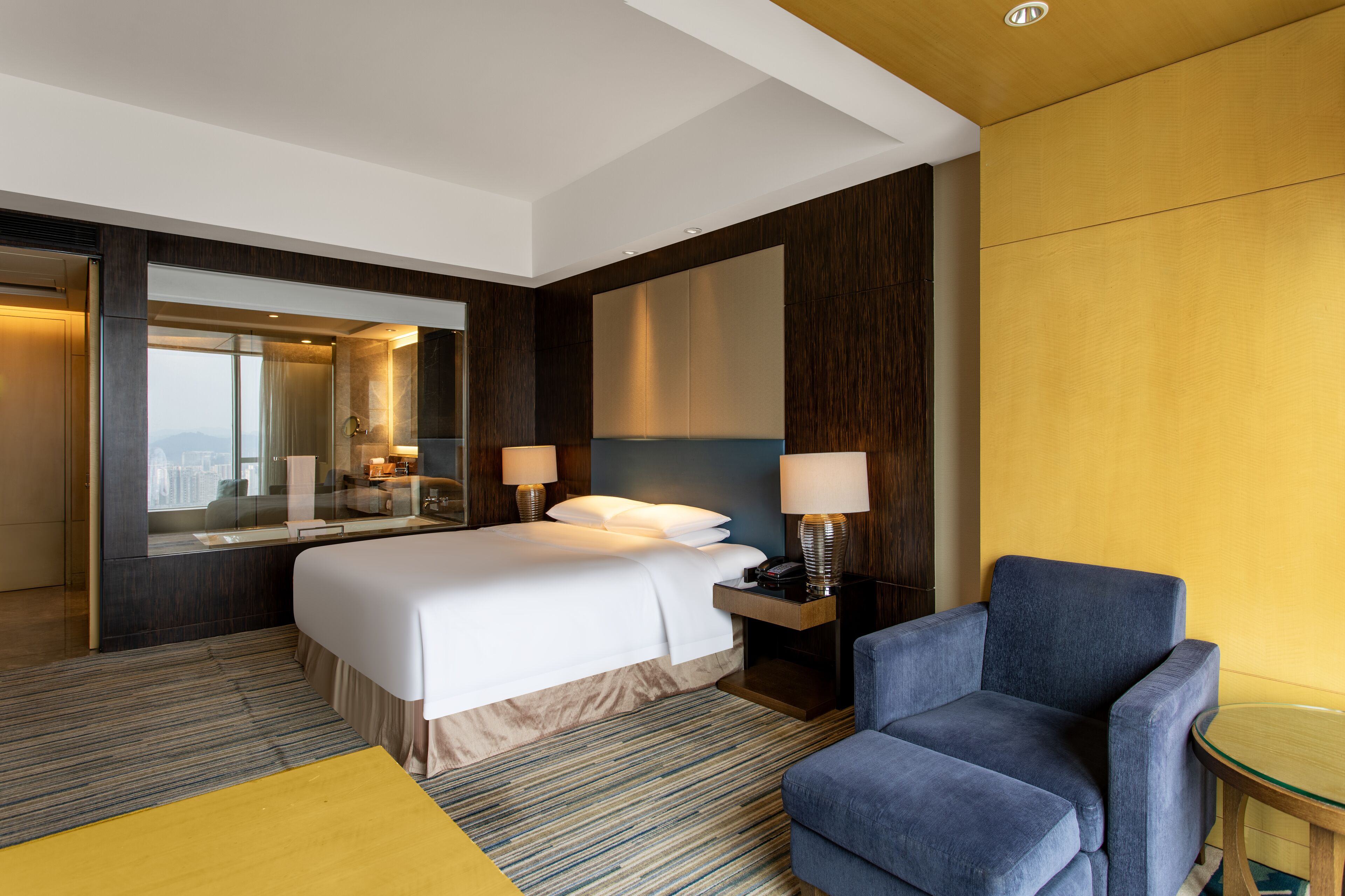 Una habitación de hotel con una cama grande, dos lámparas de noche, un sillón azul, una mesita redonda y un espejo grande.