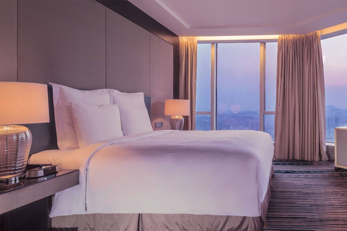 A hotel room with a large bed, a TV on a cabinet, and a city view through the window.