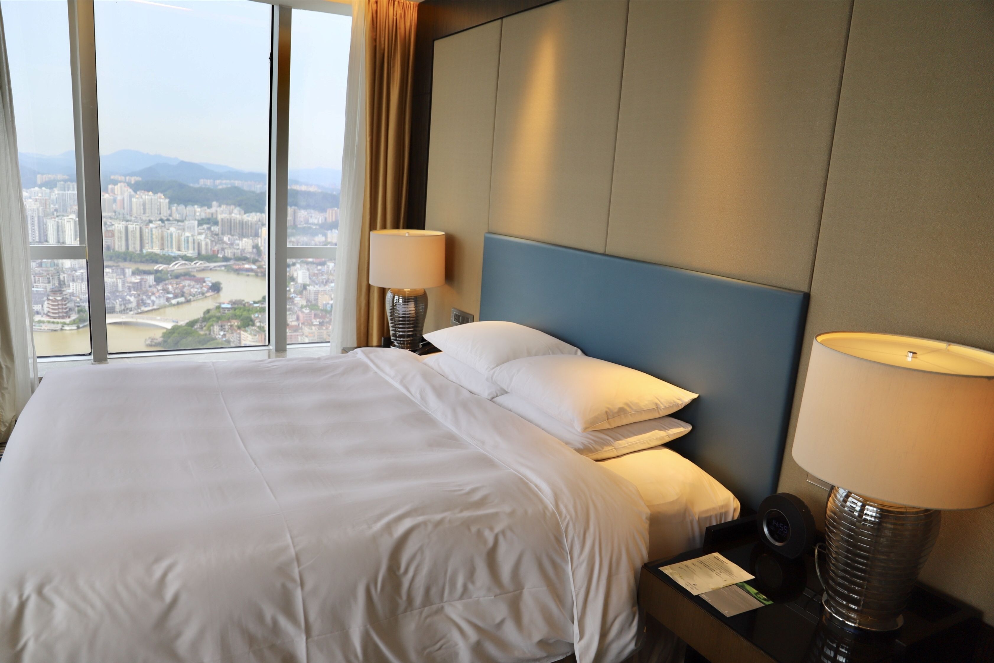 Habitación de hotel con una cama grande, dos lámparas de noche, una mesita de noche y vistas a un paisaje urbano a través de ventanas amplias.