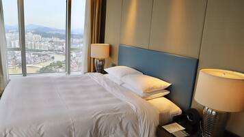 Habitación de hotel con una cama grande, dos lámparas de noche, una mesita de noche y vistas a un paisaje urbano a través de ventanas amplias.