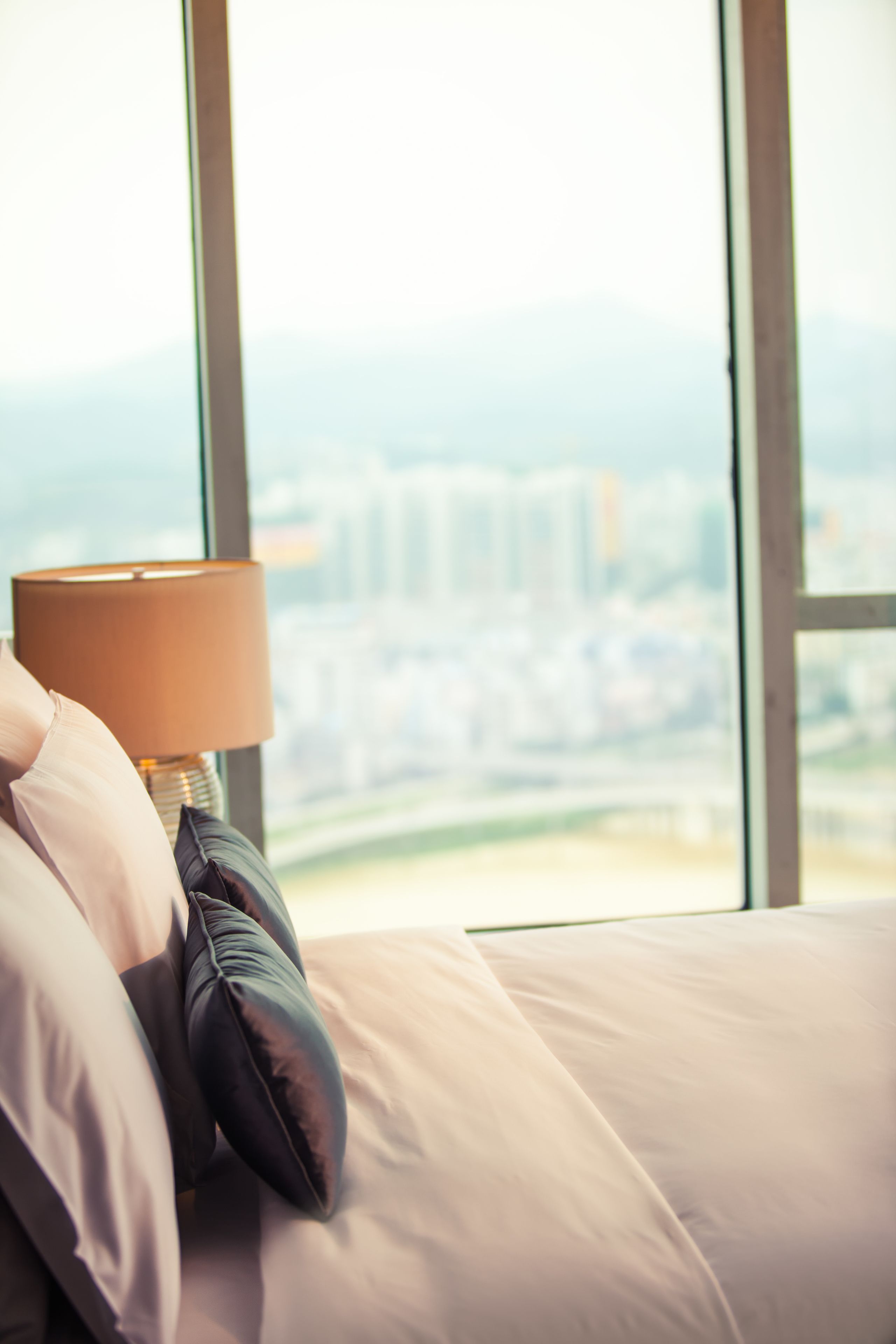Una cama con una lámpara y almohadas, un ventanal con vistas a un paisaje urbano.