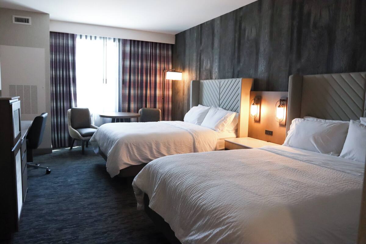 A hotel room with two beds, a desk, a chair, and a large window with curtains.