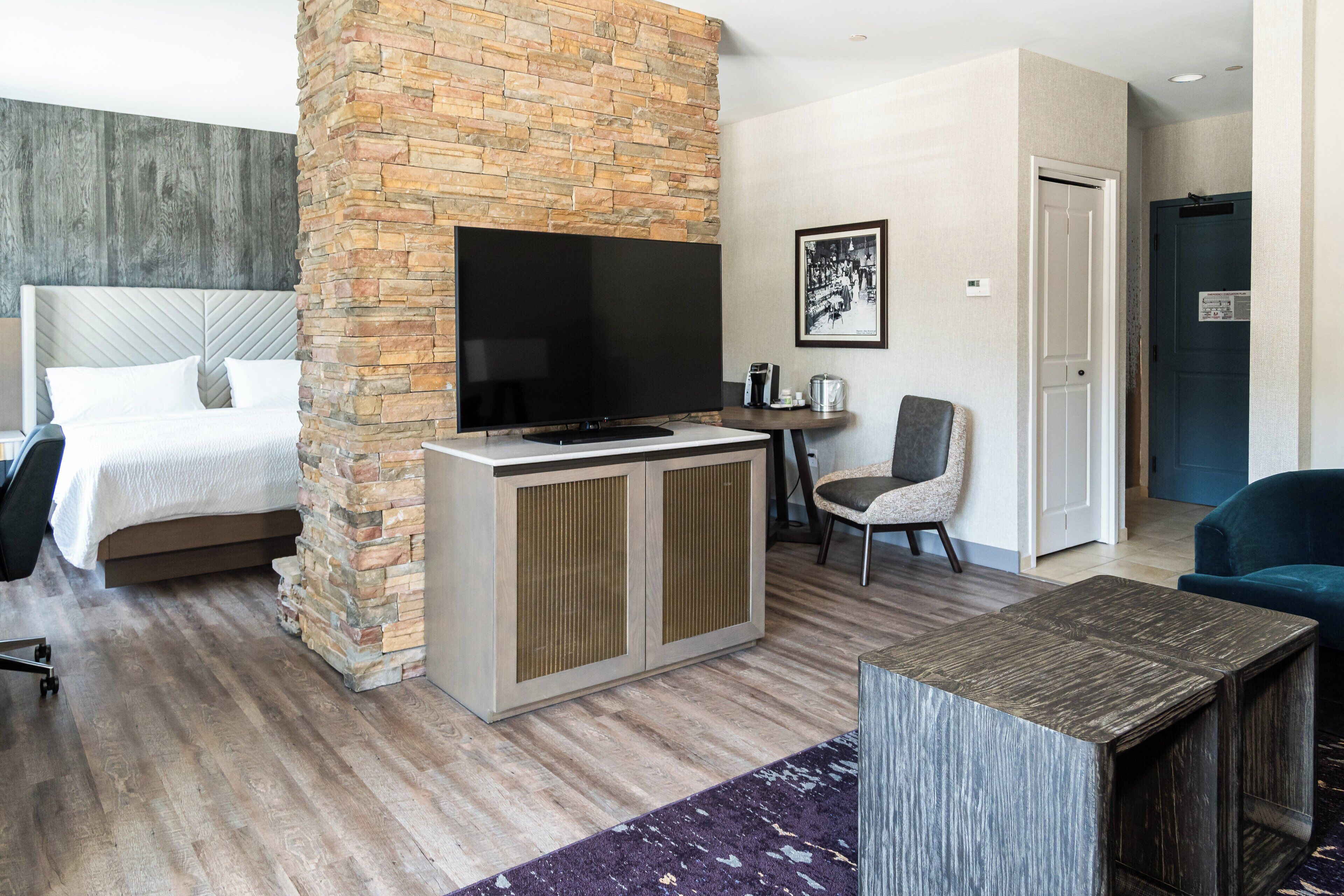 Una habitación de hotel moderna con una cama grande, una pared decorativa de piedra, un televisor de pantalla plana, una mesita, una silla y un sillón azul.
