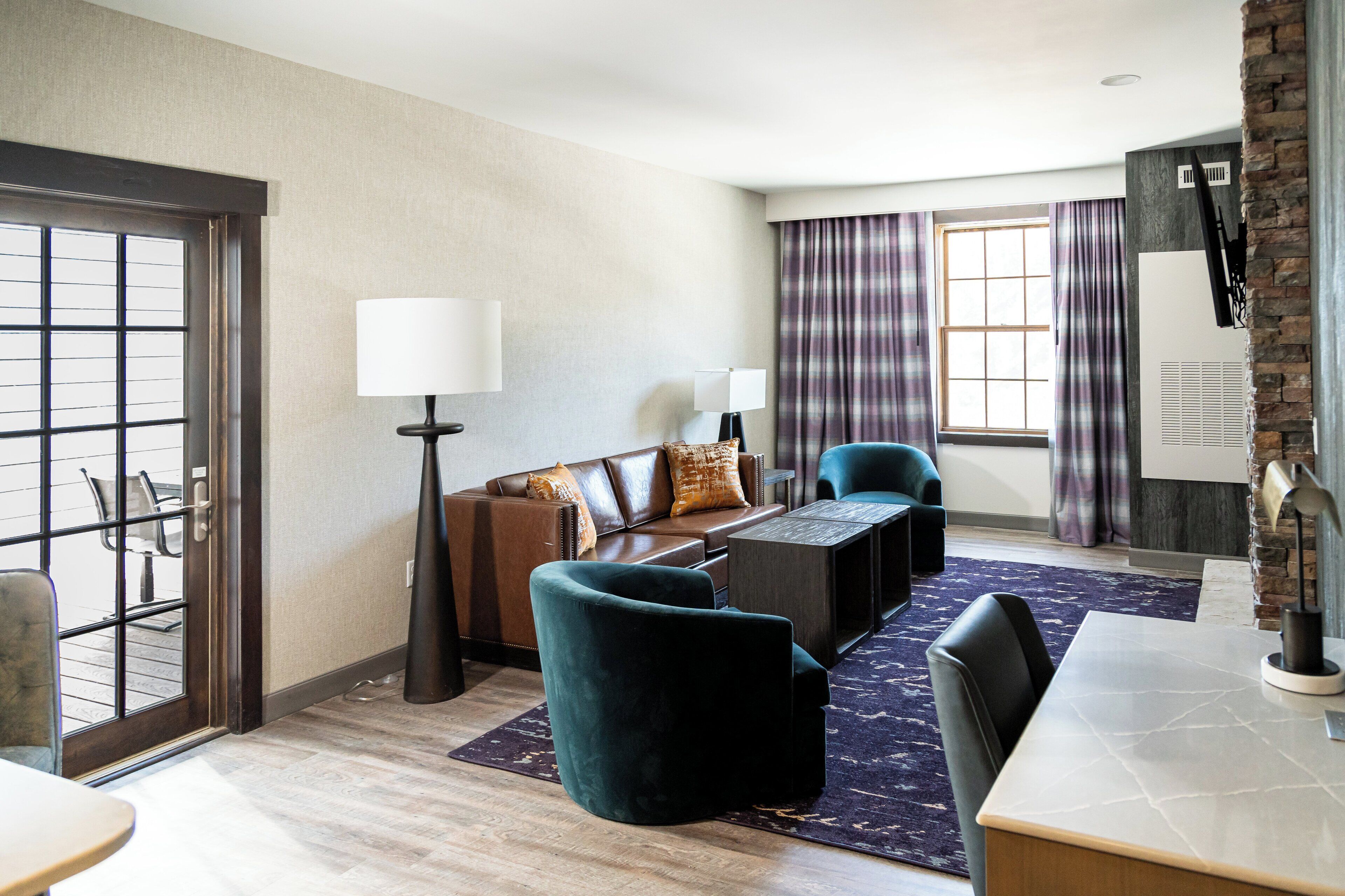 A modern hotel room with a leather sofa, two armchairs, a coffee table, and a dining table. There is a large window with curtains, a door with a view, and a stone accent wall.