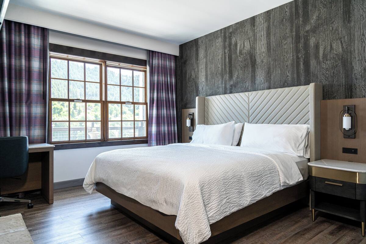 A modern hotel room with a large bed, a desk, and a view of the outdoors through a window with curtains.