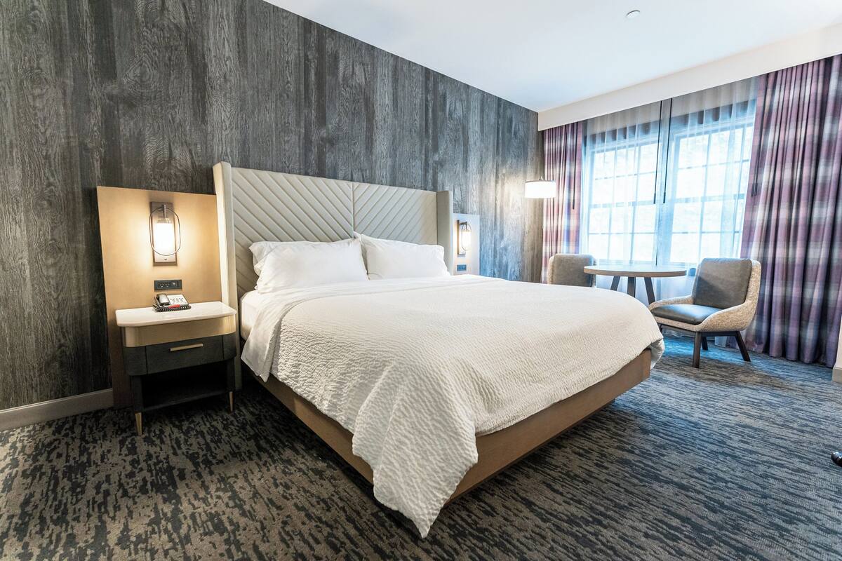 A hotel room with a large bed, a bedside table with a lamp, a chair, and a small table.