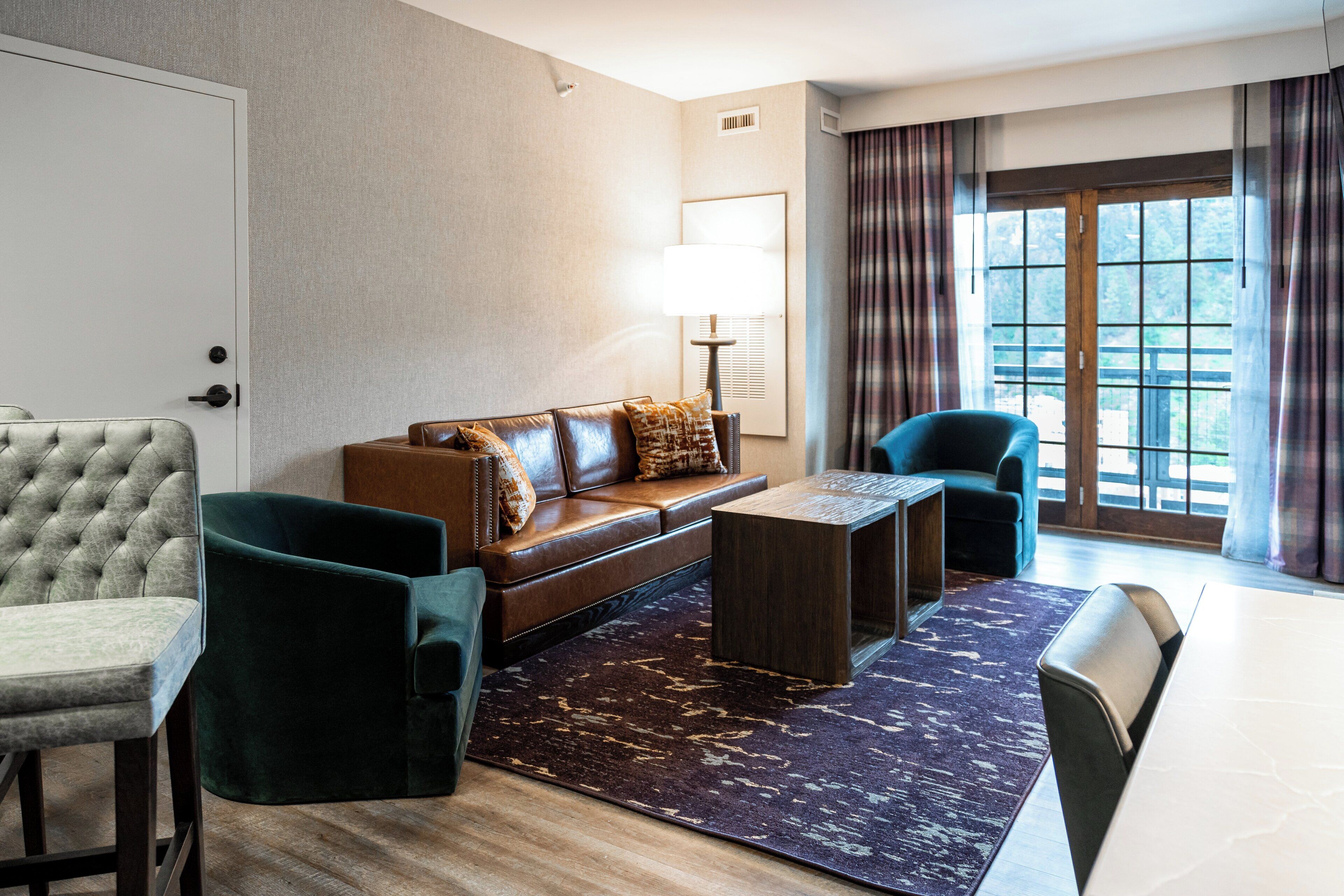 A modern hotel room with a leather sofa, two armchairs, a small coffee table, and a large window with curtains.