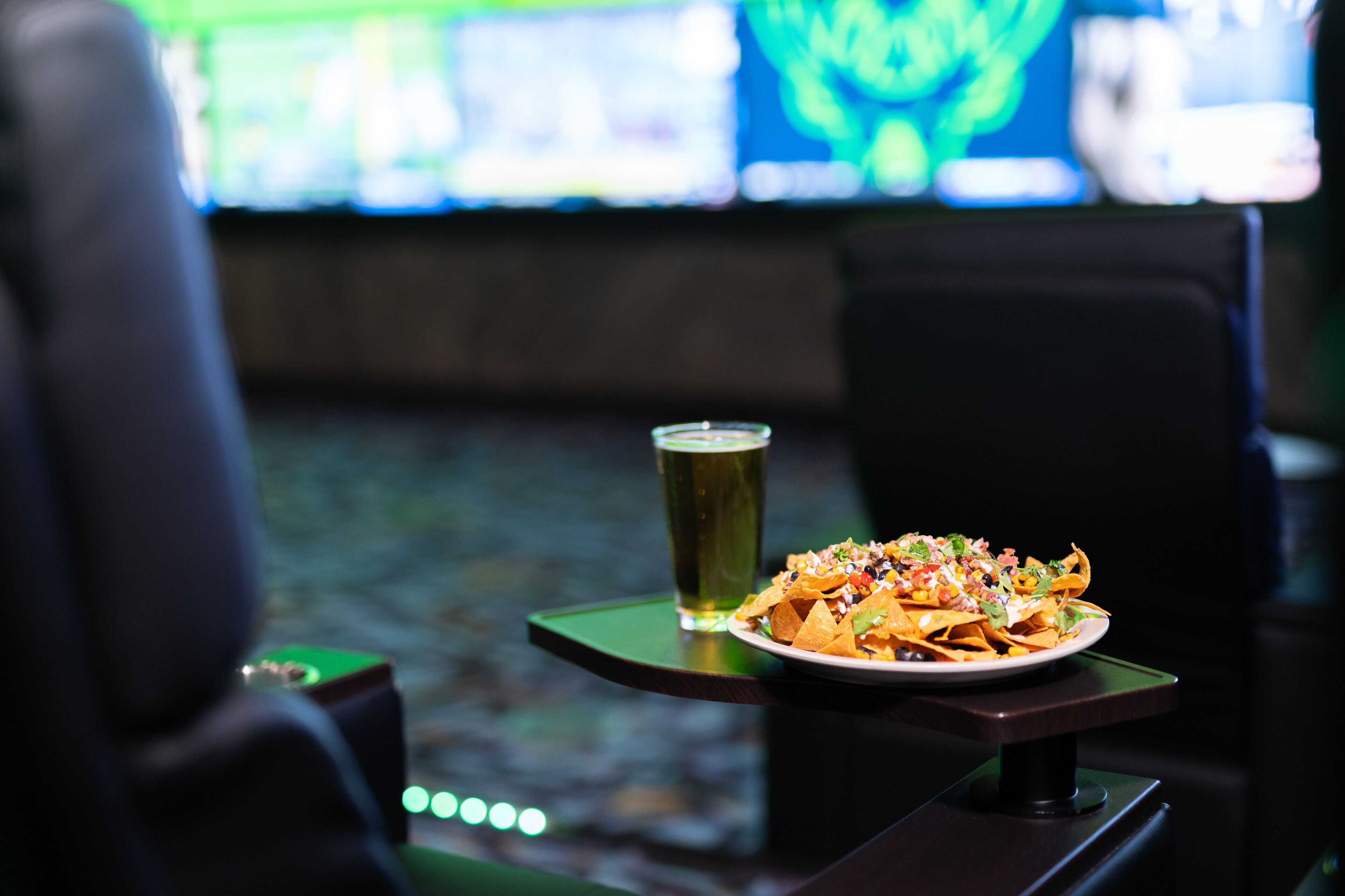 A table with nachos and a beer in a dimly lit room with a TV in the background.