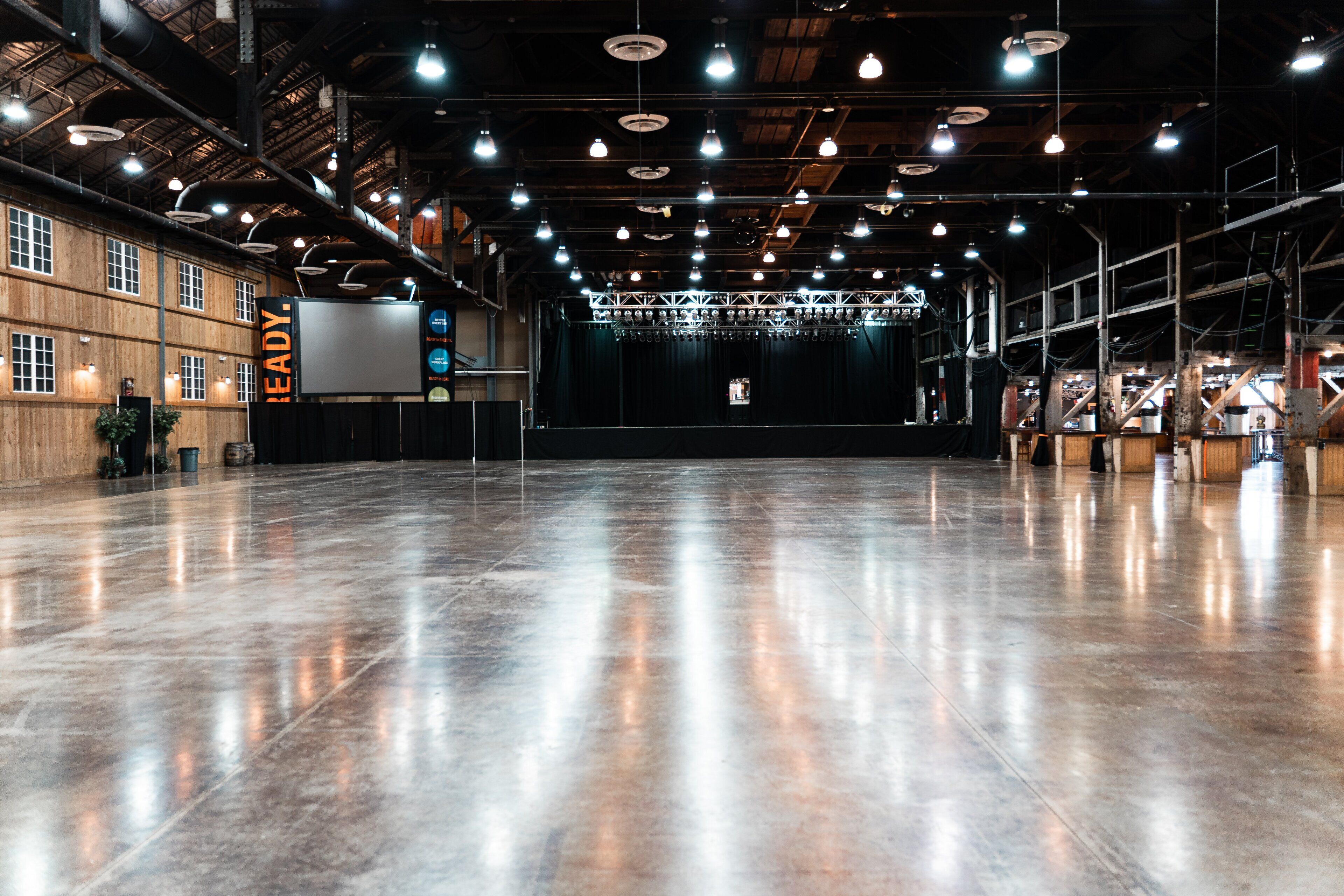 Un salón amplio de estilo industrial con suelo de concreto pulido, vigas a la vista y un escenario con cortina.