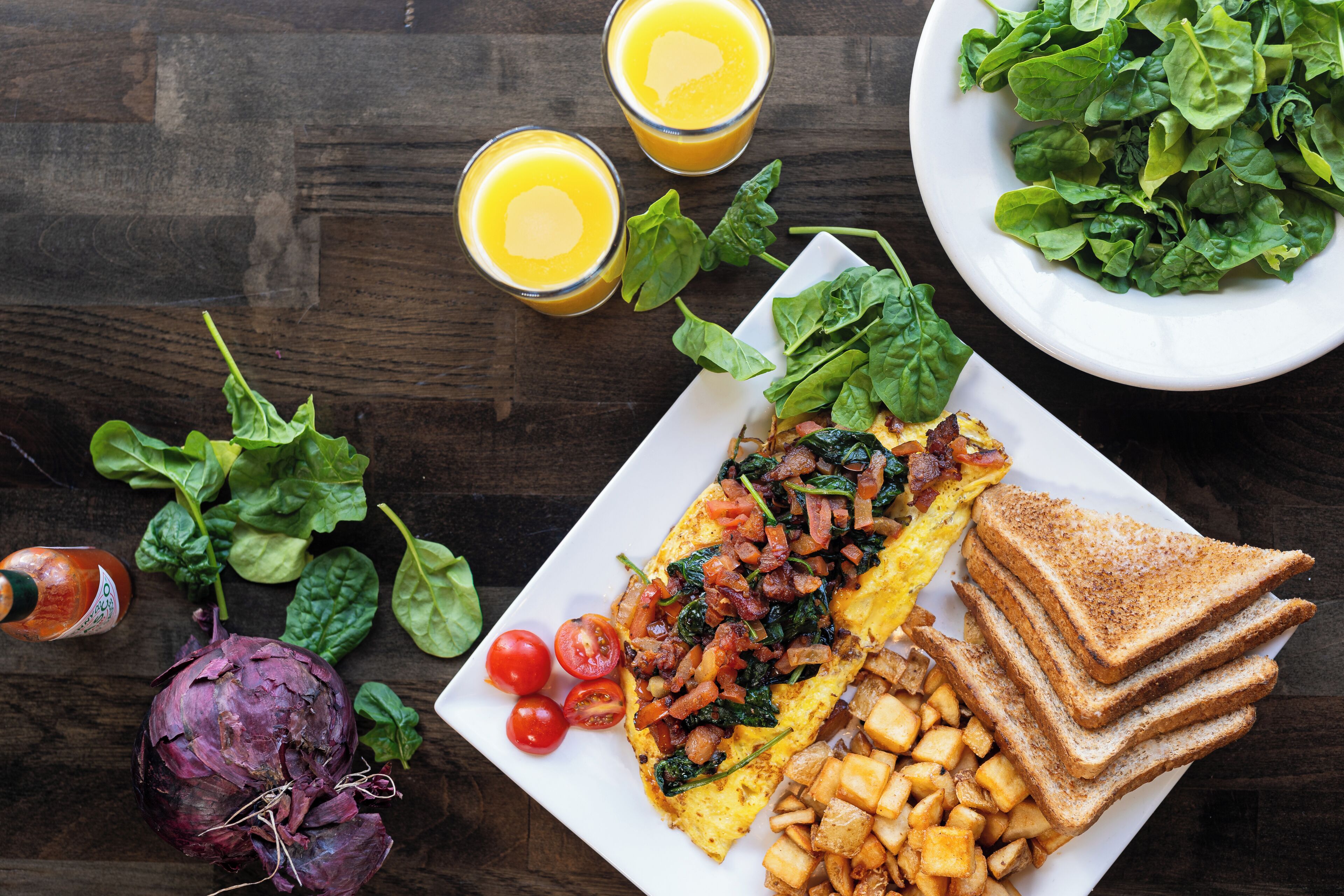 Une assiette de petit-déjeuner comprenant des toasts, des œufs et des pommes de terre, accompagnée d’épinards frais et de tomates cerises.