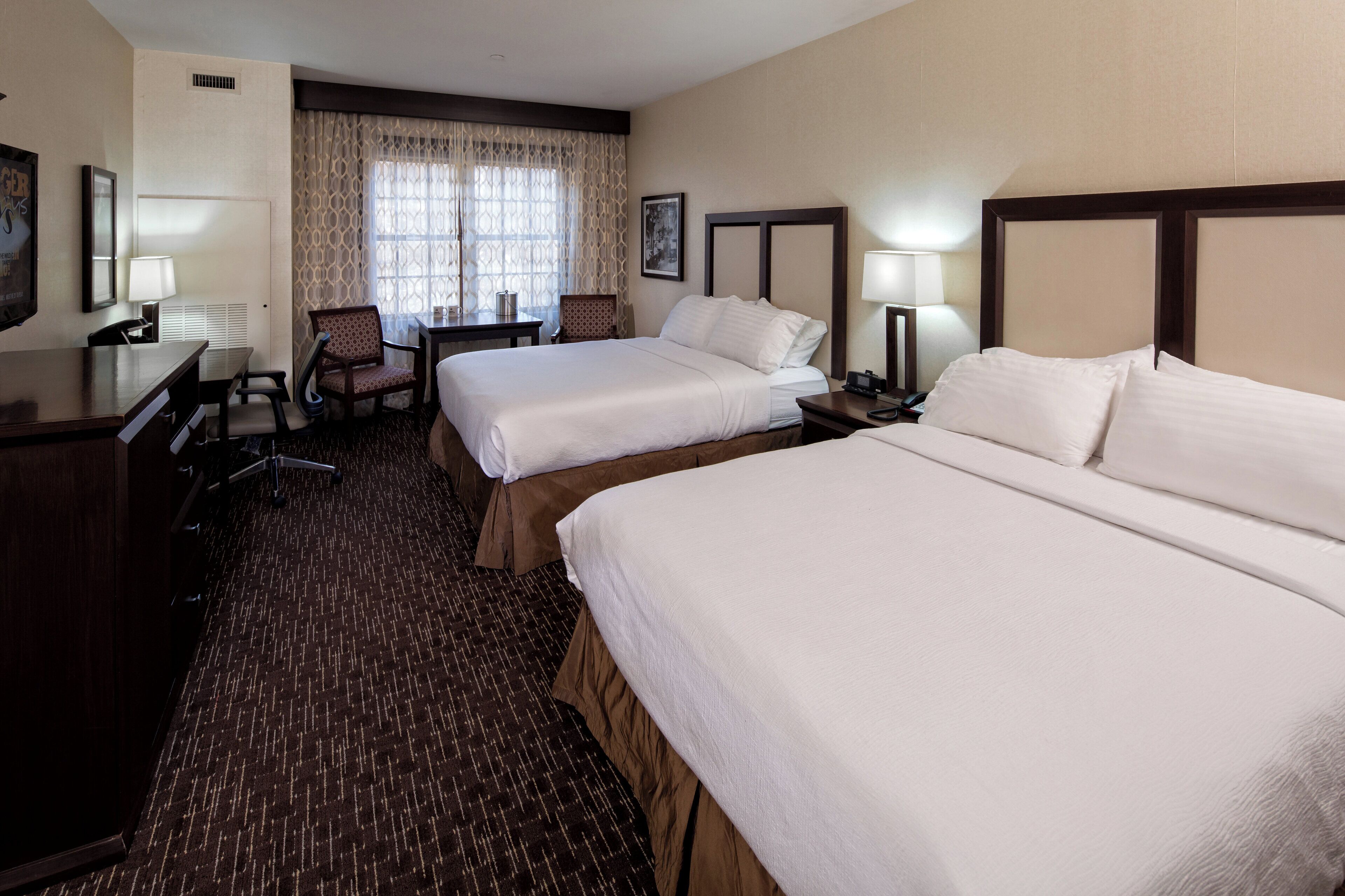 A hotel room with two beds, a desk, a chair, a lamp, and a window with curtains.