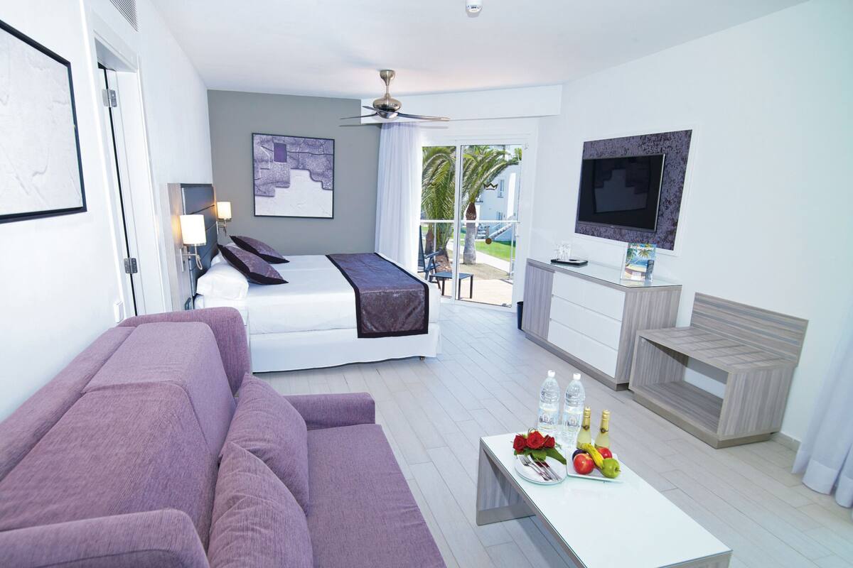 A modern hotel room with a bed, a purple sofa, a dining table with fruit and flowers, a TV, and a balcony with a view.