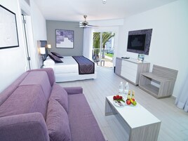 A modern hotel room with a bed, a purple sofa, a dining table with fruit and flowers, a TV, and a balcony with a view.