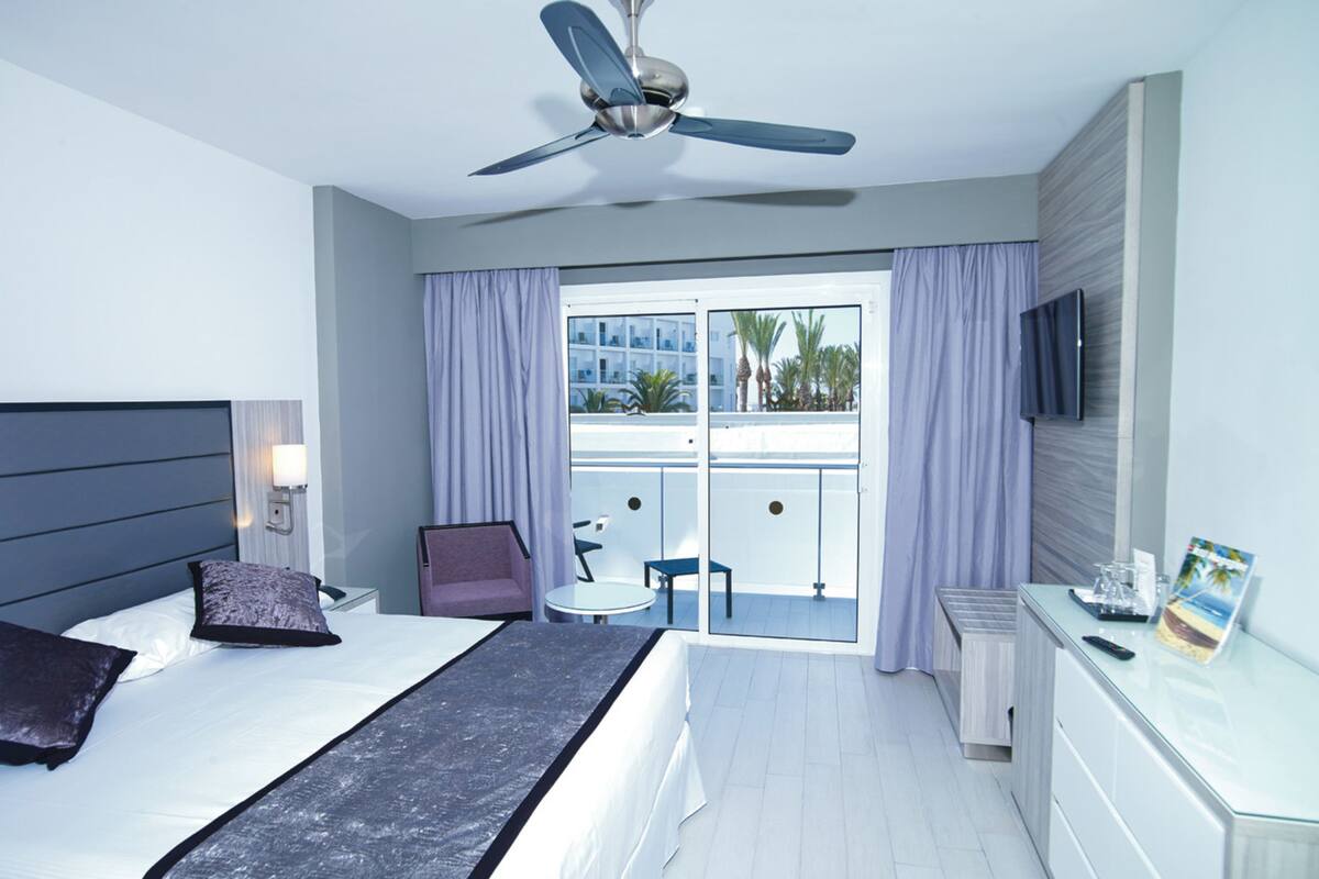 A modern hotel room with a bed, a desk, a chair, a ceiling fan, and a balcony with a view of palm trees.